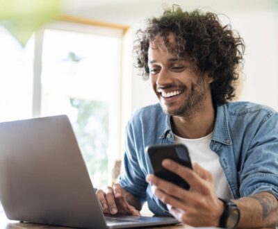 Junger Mann schaut lächelnd auf sein aufgeklapptes Laptop mit seinem Smartphone in der Hand in unserem E-Commerce Expert Kurs.