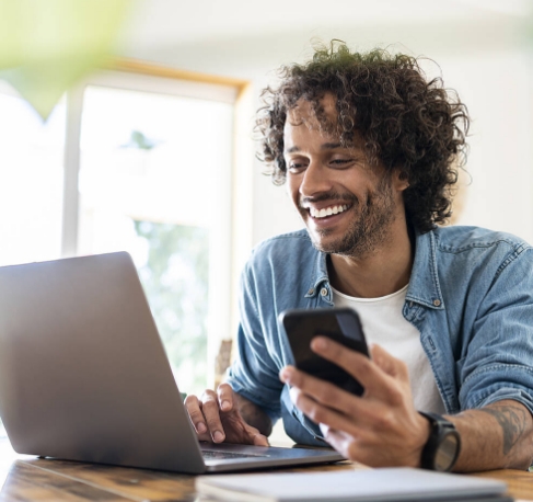 Junger Mann schaut lächelnd auf sein aufgeklapptes Laptop mit seinem Smartphone in der Hand in unserem E-Commerce Expert Kurs.