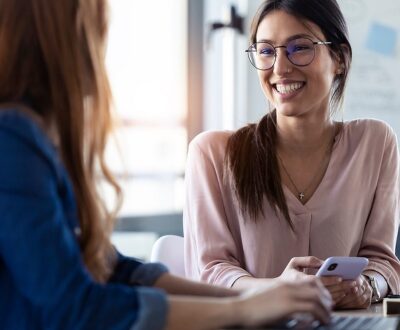 Zwei Frauen sitzen sich mit Laptop und Handy gegenüber am Schreibtisch in einer Coachingsituation