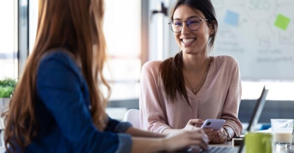 Zwei Frauen sitzen sich mit Laptop und Handy gegenüber am Schreibtisch in einer Coachingsituation