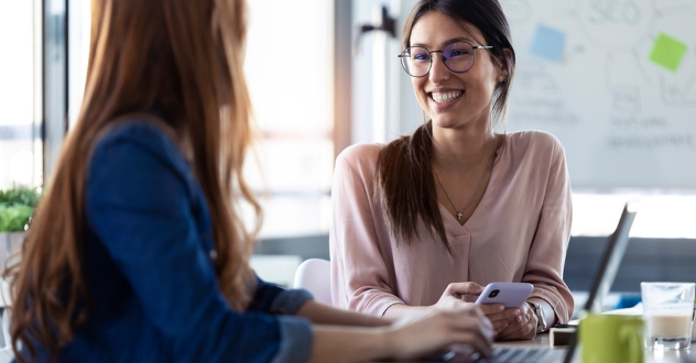 Zwei Frauen sitzen sich mit Laptop und Handy gegenüber am Schreibtisch in einer Coachingsituation