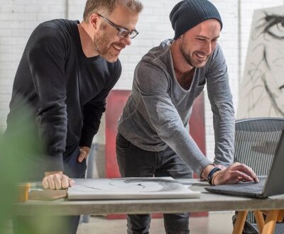 Zwei Männder schauen stehend auf einen Laptop und lächeln in unserem Gründercoaching für Designer & Künstler