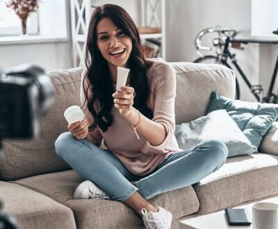 Eine Frau filmt sich beim zeigen von Kosmetikprodukten währen sie auf einem Sofa sitzt.