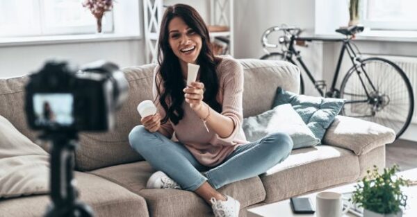 Eine Frau filmt sich beim zeigen von Kosmetikprodukten währen sie auf einem Sofa sitzt.