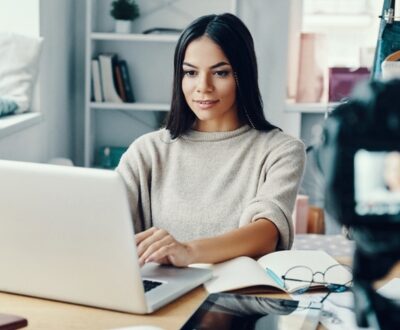 Eine Frau sitzt am Laptop und mach dabei eine Aufnahme mit einer Videokamera als Social Media Manager