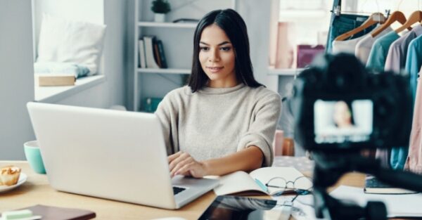 Eine Frau sitzt am Laptop und mach dabei eine Aufnahme mit einer Videokamera als Social Media Manager