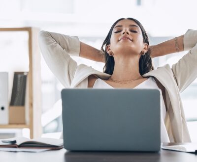 Eine Frau am Laptop lehnt sich entspannt mit hinter dem Kopf verschränkten Armen zurück in unserem Stressmanagement Coaching