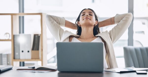 Eine Frau am Laptop lehnt sich entspannt mit hinter dem Kopf verschränkten Armen zurück in unserem Stressmanagement Coaching