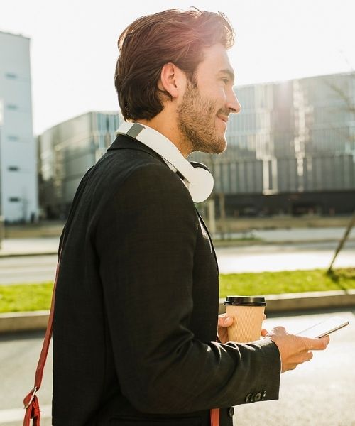 Junger Mann spaziert mit Handy und Kaffeebecher und schaut zuversichtlich auf seinen Weg in unserem Business Coaching