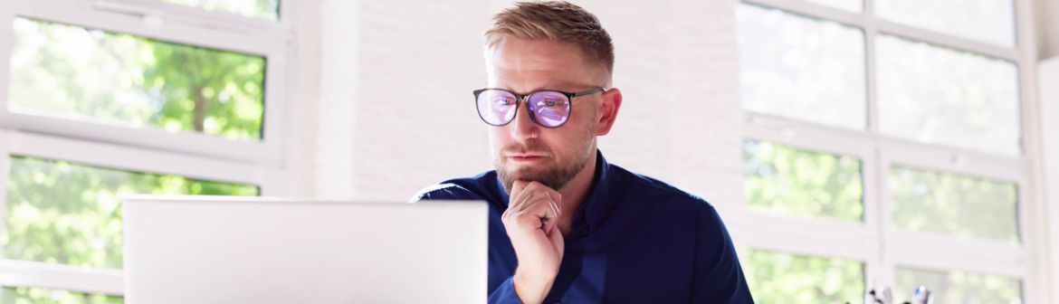 Arbeitslos – was nun? Ein Mann mit Brille und dunkelblauem Hemd sitzt konzentriert vor einem Laptop in einem hellen Büro mit großen Fenstern und denkt über seine nächsten Schritte nach.