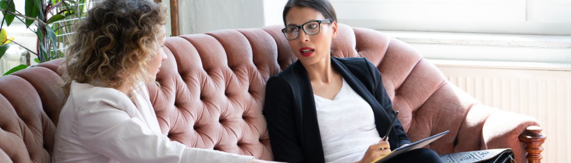 Systemisches Coaching: Zwei Frauen sitzen auf einem rosa Sofa in einem Beratungsgespräch. Die Coachin hält Notizen in der Hand, während die Klientin aufmerksam zuhört