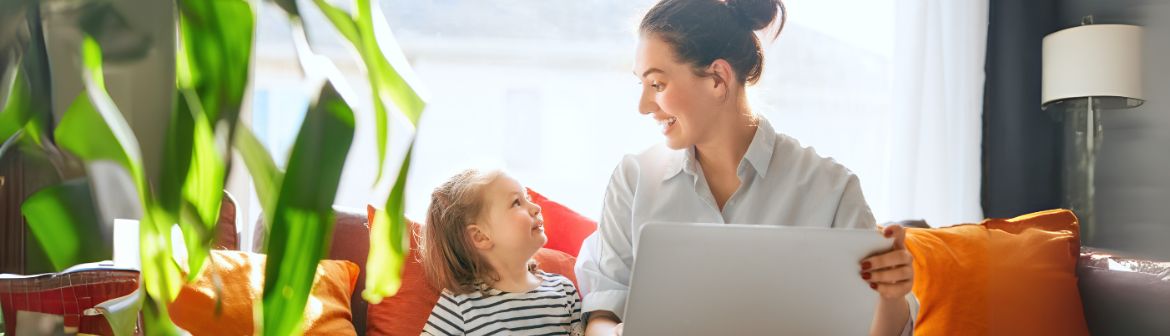 Weiterbildung in der Elternzeit: Eine lächelnde Mutter mit Laptop sitzt auf einem gemütlichen Sofa und schaut ihr Kind an, das neben ihr sitzt. Die perfekte Balance zwischen Familie und beruflicher Weiterentwicklung