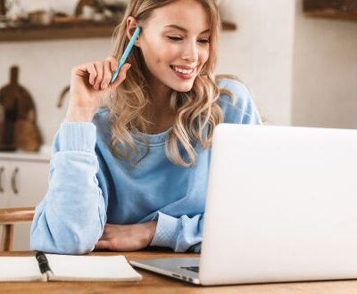 Junge Frau in gemütlicher Umgebung bei einer Live-Online-Weiterbildung, mit Laptop und Notizbuch am Holztisch.