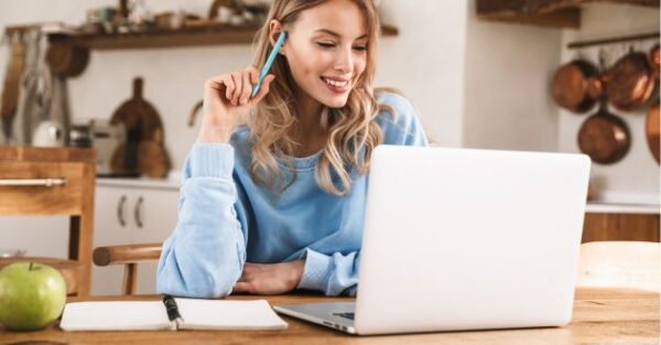 Junge Frau in gemütlicher Umgebung bei einer Live-Online-Weiterbildung, mit Laptop und Notizbuch am Holztisch.