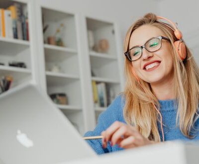 Junge Frau mit Kopfhörern und Laptop in einer Live-Online-Weiterbildung, lachend und konzentriert in ihrem Arbeitszimmer.