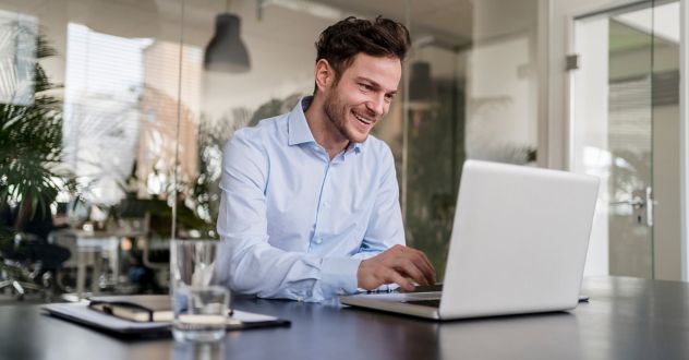 Ein junger Mann in Business-Kleidung sitzt in einem modernen Büro und arbeitet konzentriert an seinem Laptop. Er nimmt an einer Weiterbildung zur KI-Anwendung und Implementierung teil, während er lächelnd auf den Bildschirm schaut. Das Bild vermittelt digitale Lernprozesse, berufliche Weiterbildung und den praktischen Einsatz von Künstlicher Intelligenz im Unternehmensumfeld.