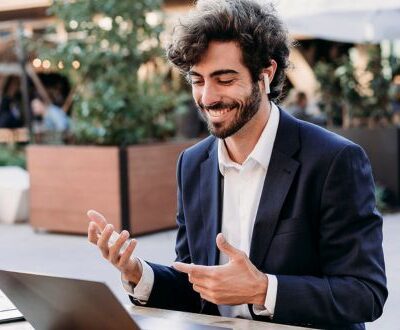 Ein junger Mann in Business-Kleidung nimmt mit kabellosen Kopfhörern an einer Online-Weiterbildung zu KI-Implementierung und agilen Methoden teil. Er sitzt draußen an einem Laptop und führt eine Videokonferenz mit einem Lächeln, während er gestikuliert.