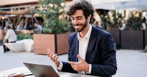 Ein junger Mann in Business-Kleidung nimmt mit kabellosen Kopfhörern an einer Online-Weiterbildung zu KI-Implementierung und agilen Methoden teil. Er sitzt draußen an einem Laptop und führt eine Videokonferenz mit einem Lächeln, während er gestikuliert.