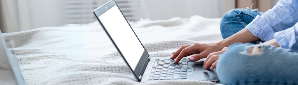 Eine Frau sitzt auf dem Bett und tippt auf ihrem geöffneten Laptop.